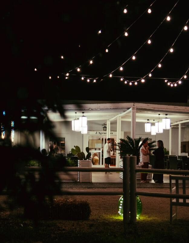 Locale serale all'aperto con luci sospese e persone che conversano.