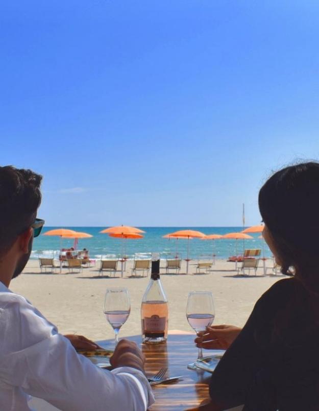 Coppia al mare con vino, ombrelloni arancioni e cielo limpido.