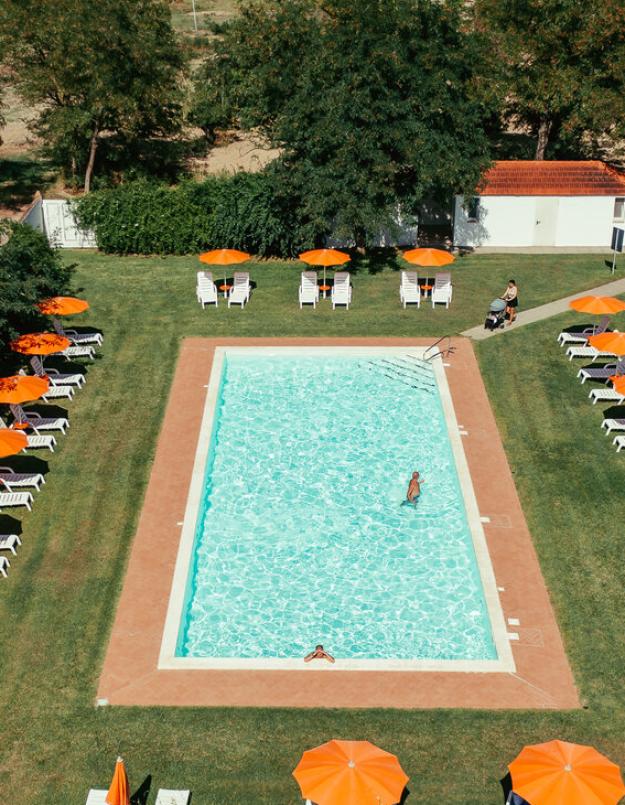 Piscina all'aperto con lettini e ombrelloni arancioni, circondata da alberi e prato.