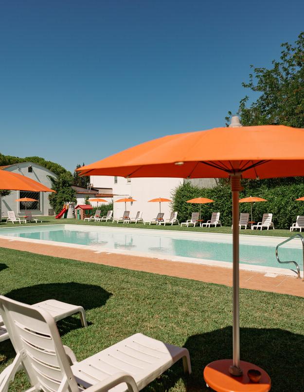 Piscina all'aperto con ombrelloni arancioni e lettini su prato verde.