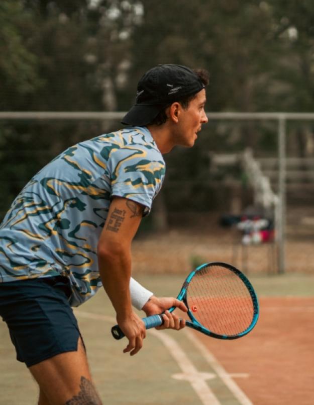 Giocatore di tennis in attesa, con racchetta blu su campo all'aperto.