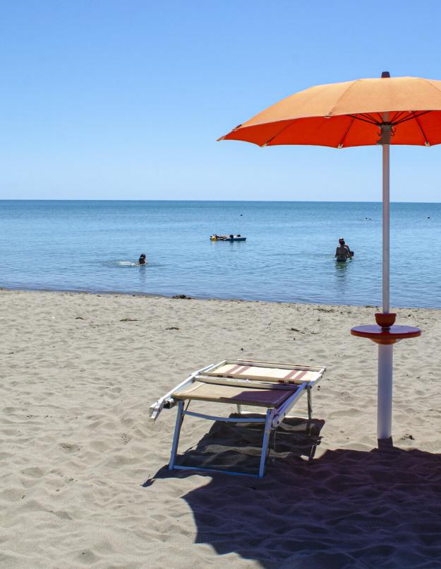 Spiaggia sabbiosa con ombrellone arancione, sdraio e persone che nuotano nel mare.