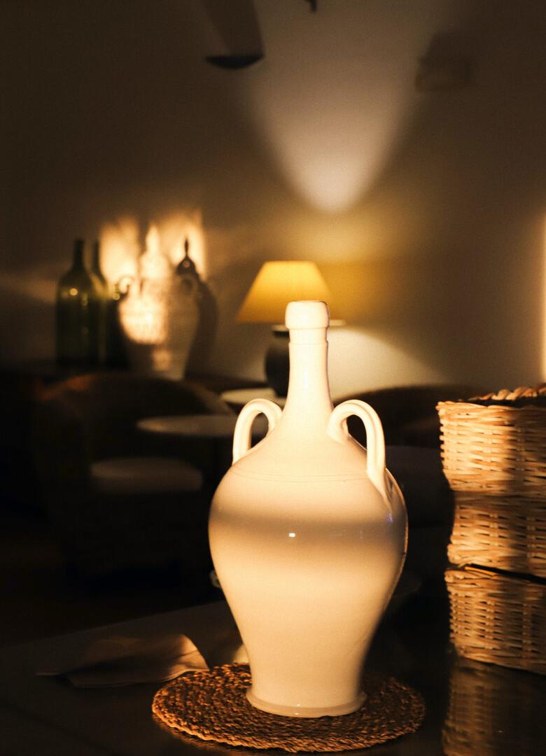 Anfora bianca su tavolo, illuminata da lampada, in ambiente accogliente e caldo.